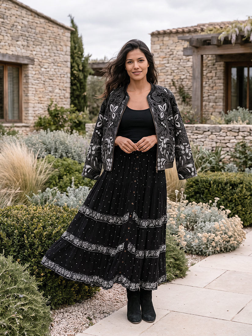 A woman stands outdoors in a garden courtyard wearing a black maxi dress with silver embroidered details, a matching black top, and a black jacket with silver floral embroidery. Stone buildings and green plants are visible in the background.