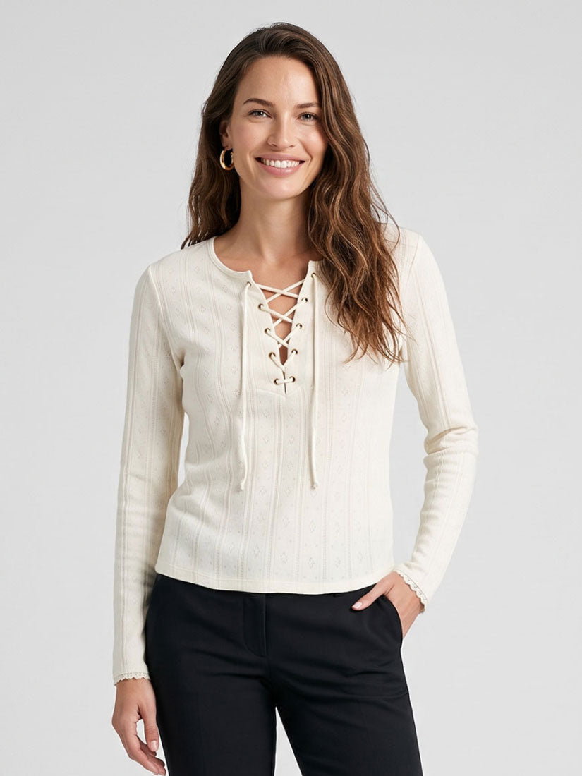 A woman modeling a cream ribbed knit long-sleeve top with a lace-up front, paired with black pants, against a light gray studio background.