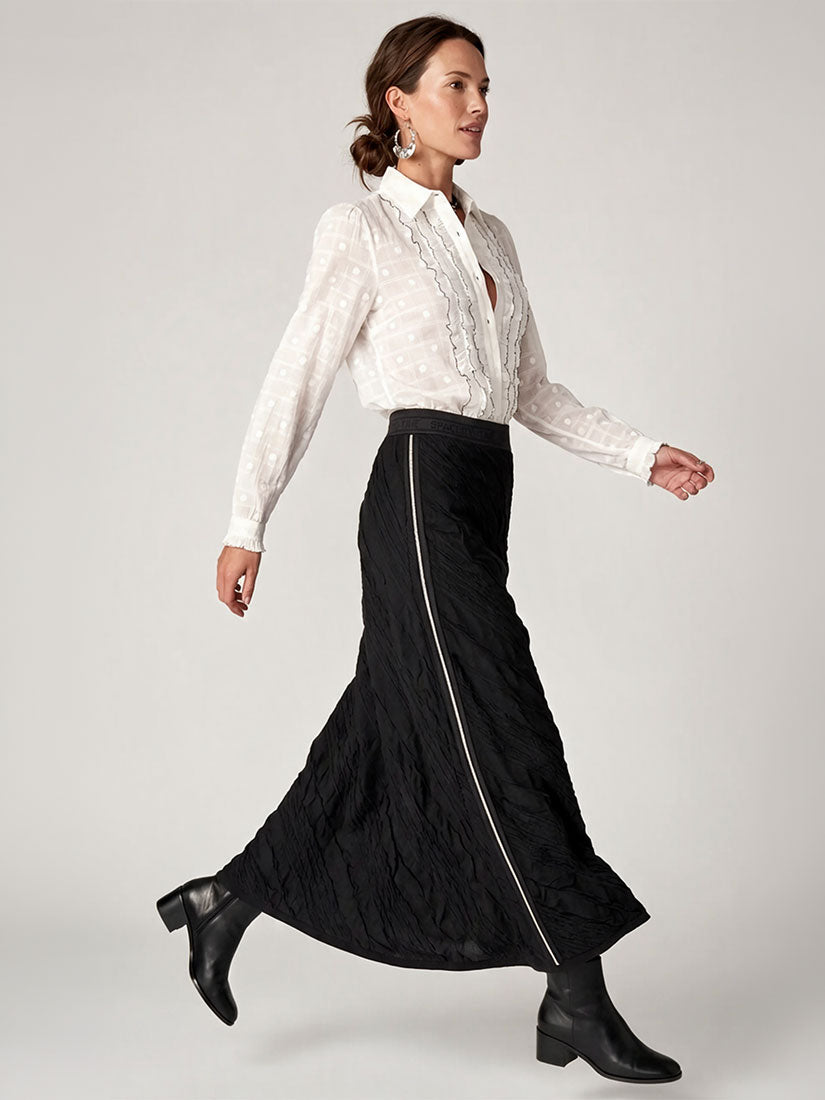 A woman modeling a long black quilted skirt with a silver side zipper, paired with a white lace-trim blouse and black ankle boots, against a light gray studio background.