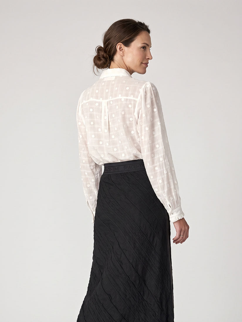 A woman shown from the back wearing a sheer white long-sleeve blouse with a subtle geometric pattern and a black textured midi skirt, photographed against a light gray studio background.