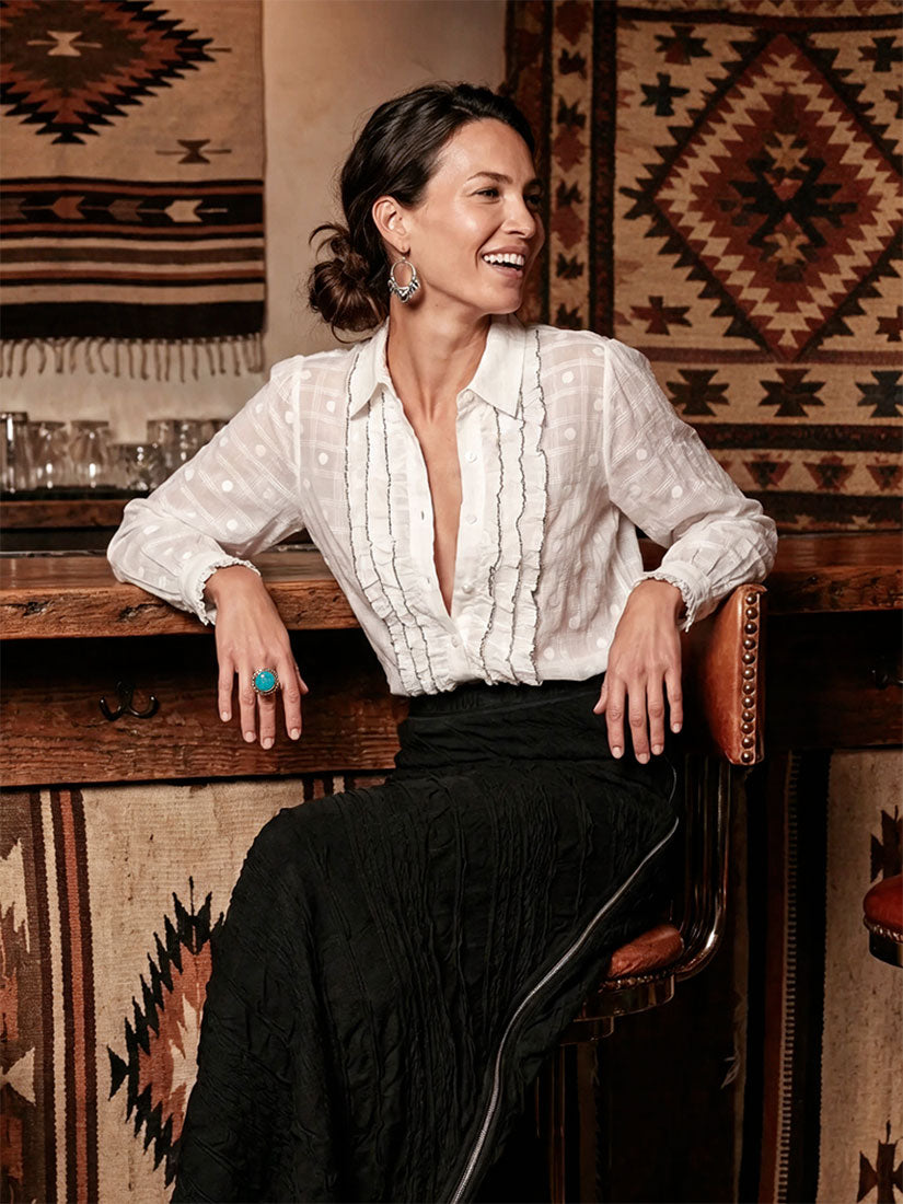 A woman smiling while seated on a wooden stool in a rustic interior, wearing a white long-sleeve blouse with vertical ruffle detailing and a black skirt. Patterned woven textiles hang on the wall behind her.