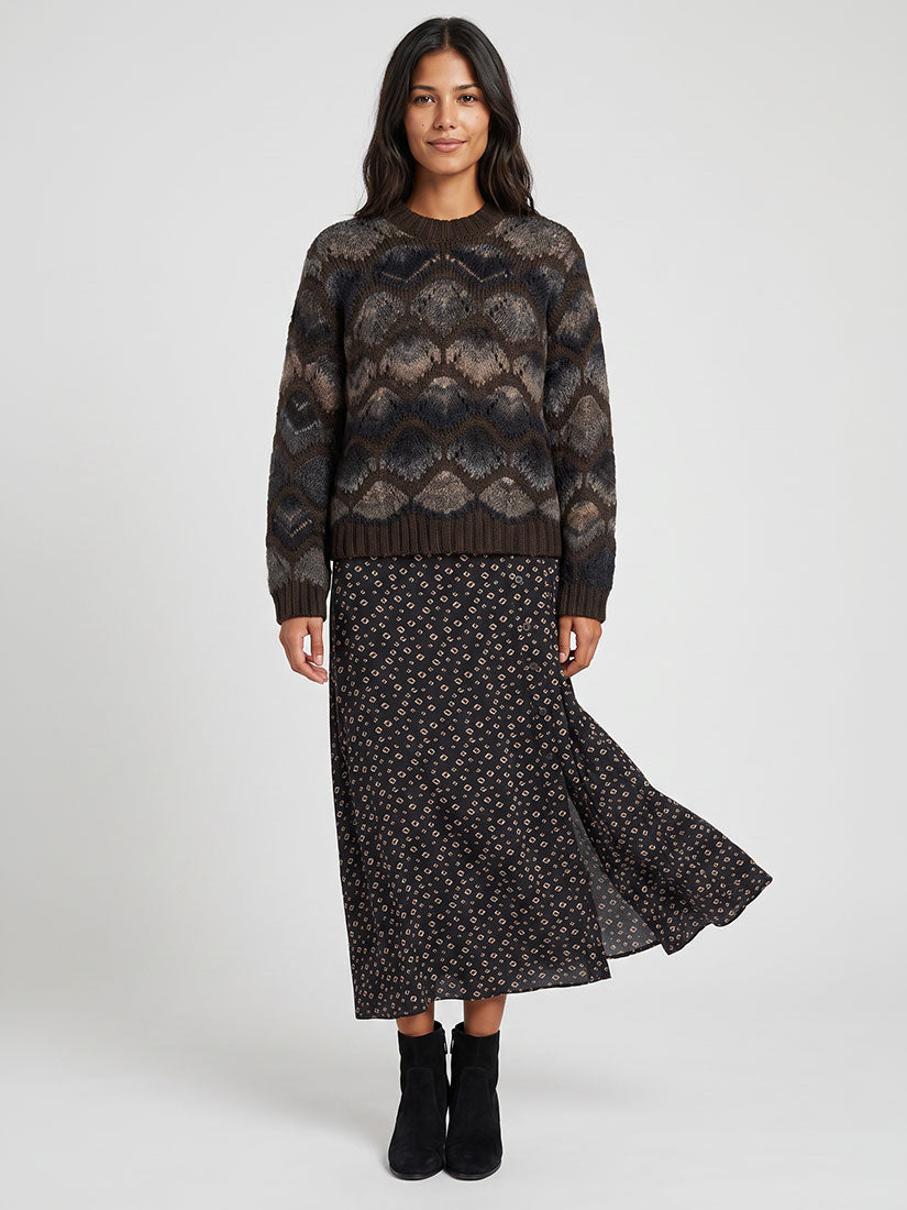 A woman modeling a dark brown patterned knit sweater and a matching dark floral midi skirt with black ankle boots, shot against a light gray studio background.