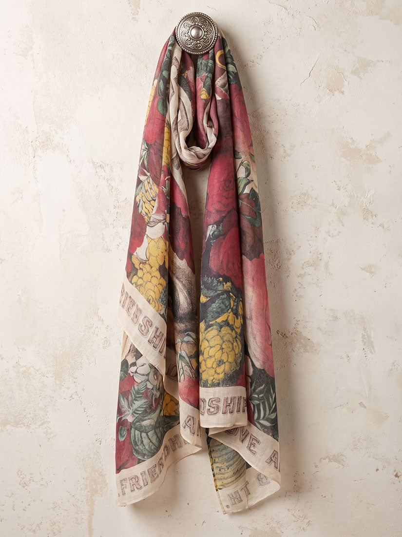 A lightweight scarf with a vintage-style botanical and fruit print in muted red, yellow, green, and beige tones, draped over a round silver wall hook against a textured beige plaster wall.