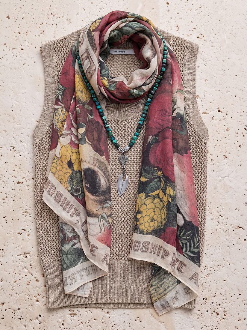 A styled flat-lay of a beige knit sweater vest with a colorful floral scarf draped over it, accessorized with a turquoise beaded necklace and a silver pendant, set against a textured beige stone background.