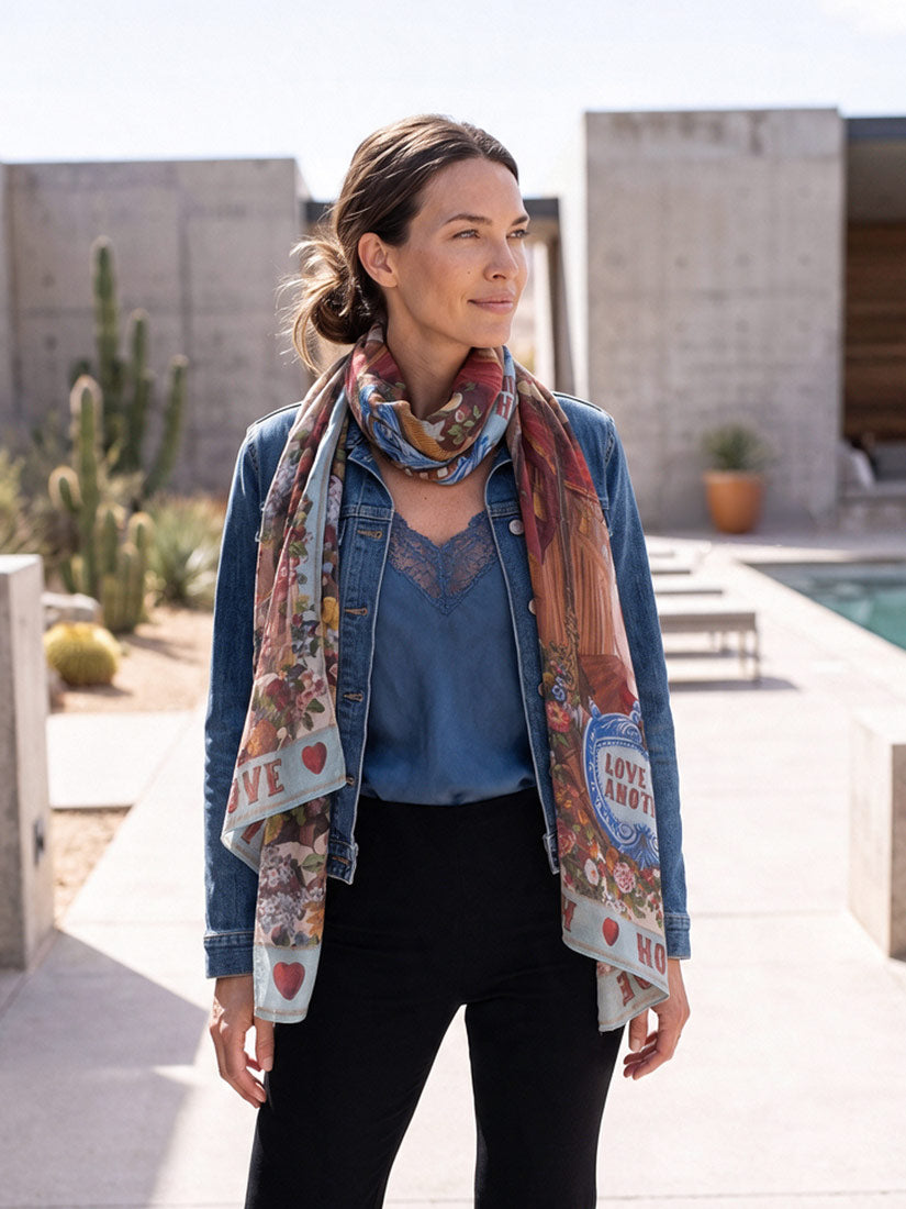 A woman wearing a blue denim jacket, blue top, black pants, and a colorful patterned scarf with hearts and text, standing outdoors in a modern concrete courtyard.