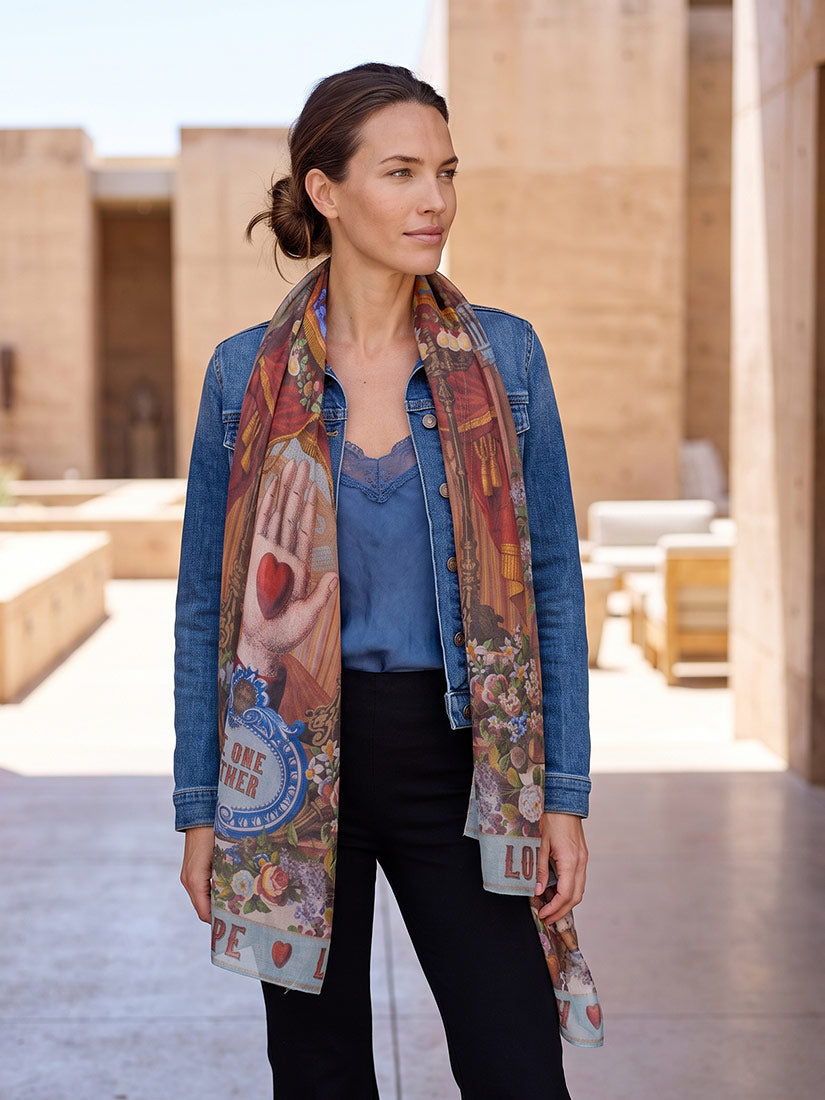 A woman wearing a blue denim jacket, blue top, black pants, and a long multicolor printed scarf with floral and graphic designs, standing in an outdoor architectural setting.