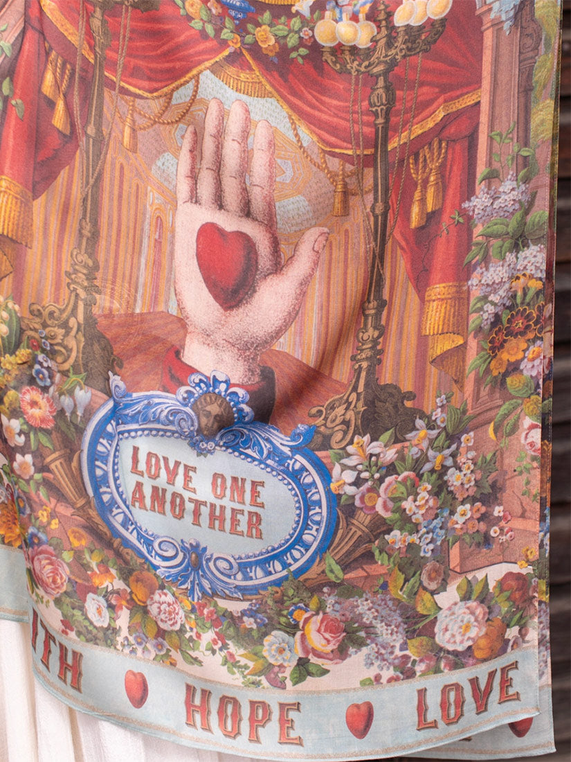 A close-up of a printed textile featuring a vintage-style illustration with a raised hand holding a red heart, red drapery, floral arrangements, and the text 'LOVE ONE ANOTHER' on a blue sign. The fabric is shown hanging, with part of a white garment or surface visible at the lower left.