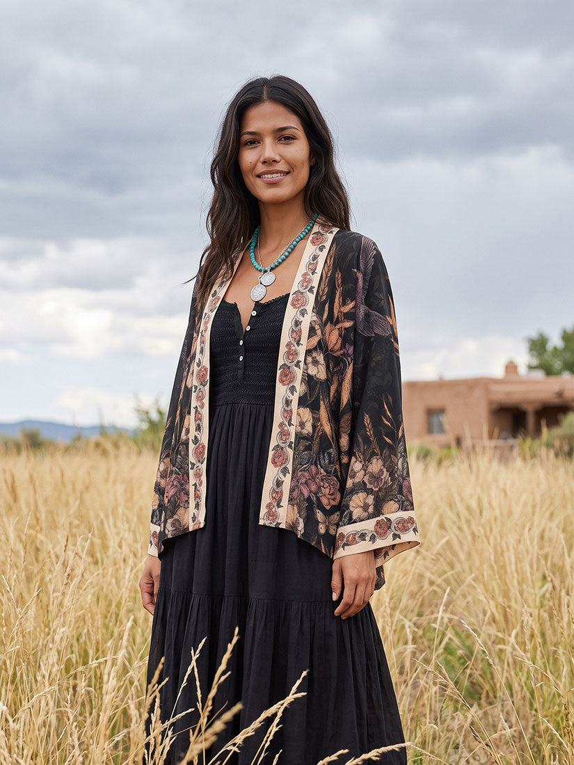 A woman stands in a grassy field wearing a black dress with a sheer floral cardigan. The cardigan has a beige border with a floral pattern and is styled with a turquoise necklace.