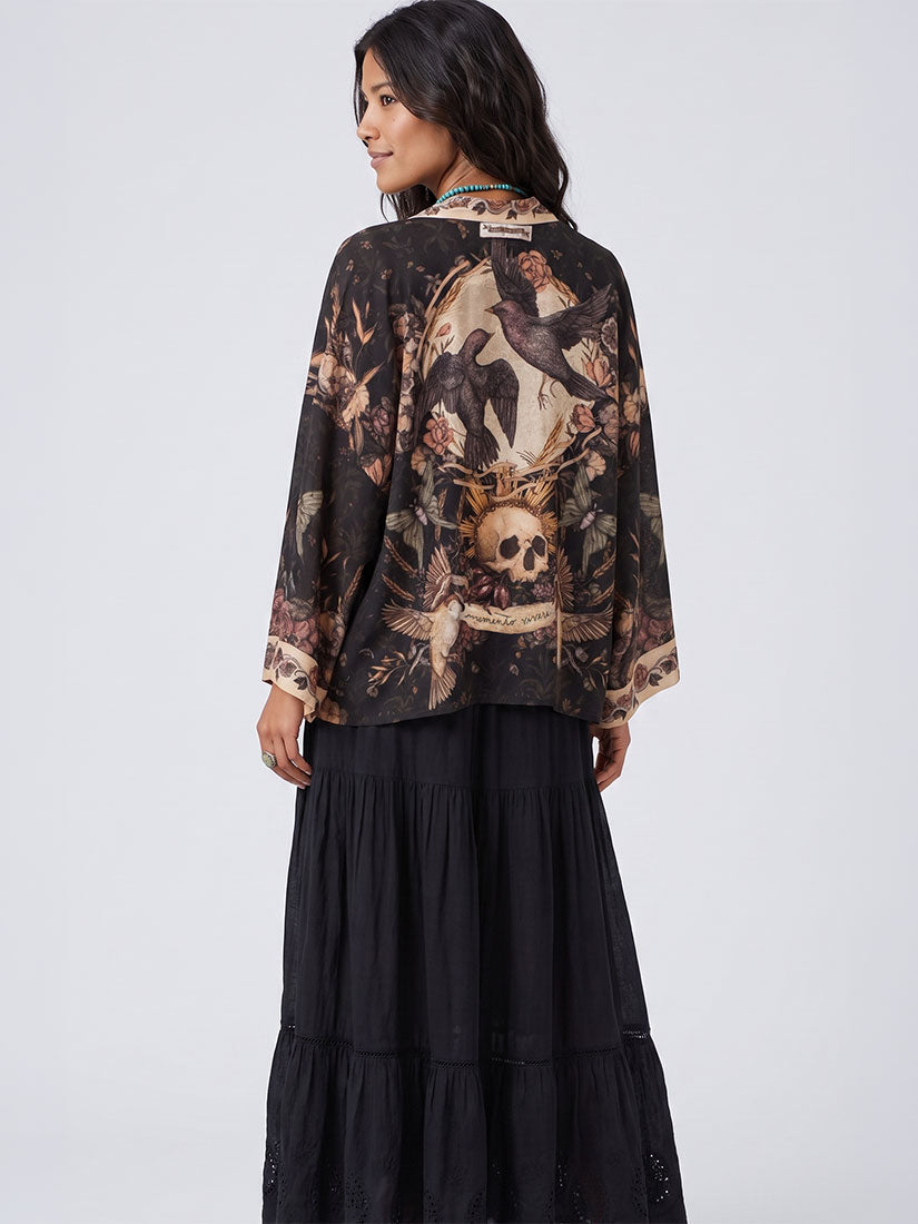 A woman shown from the back wearing a black long-sleeve blouse with a large vintage-style graphic print featuring a skull, birds, and floral motifs, paired with a long black tiered skirt, against a light gray studio background.