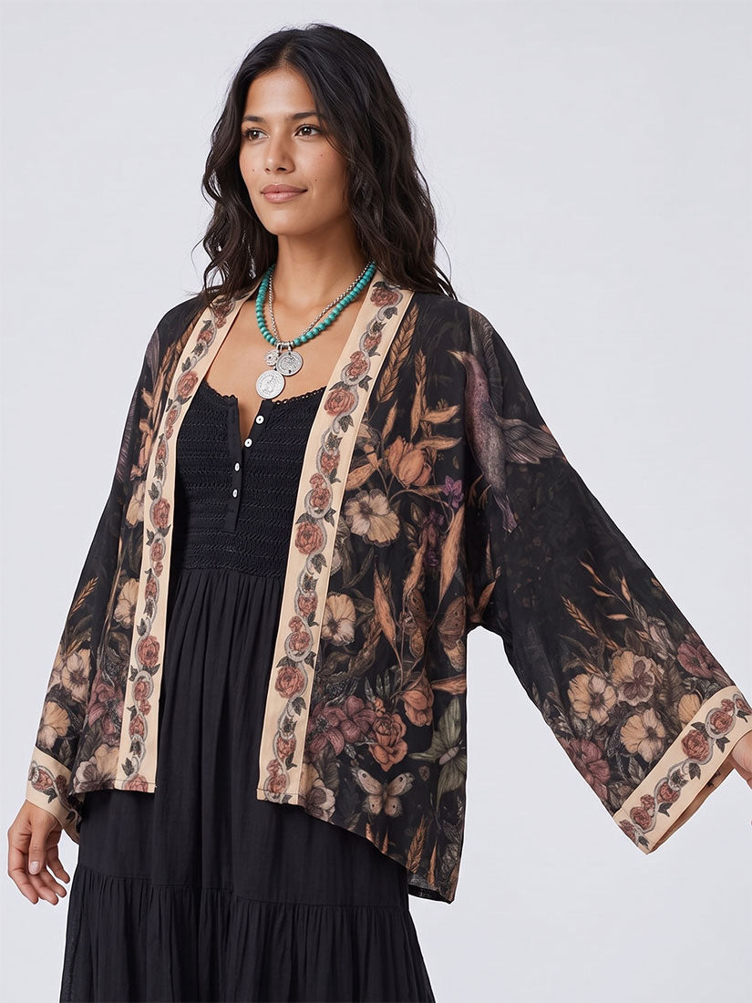 A woman wearing a black open-front kimono-style cardigan with a brown and beige floral print and wide sleeves, layered over a black dress, against a light gray studio background.
