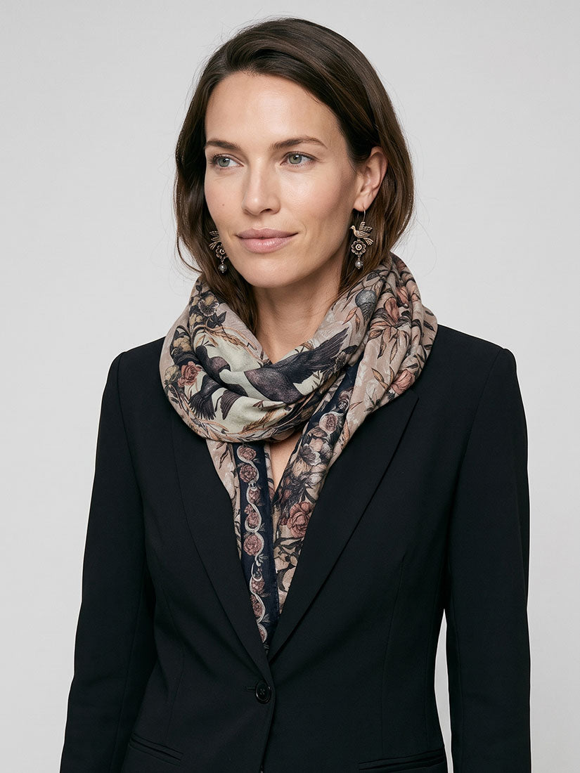 A woman wearing a black blazer and a patterned scarf with birds and floral motifs in beige, brown, and muted blue, photographed against a light gray studio background.