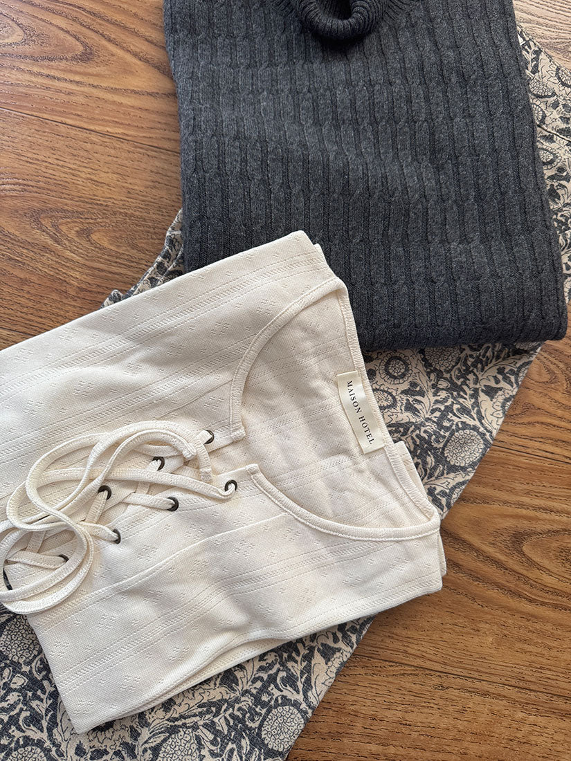 A folded cream lace-up top and a folded dark charcoal ribbed sweater are arranged on a patterned fabric over a wooden floor. A small neck label is visible on the cream top.