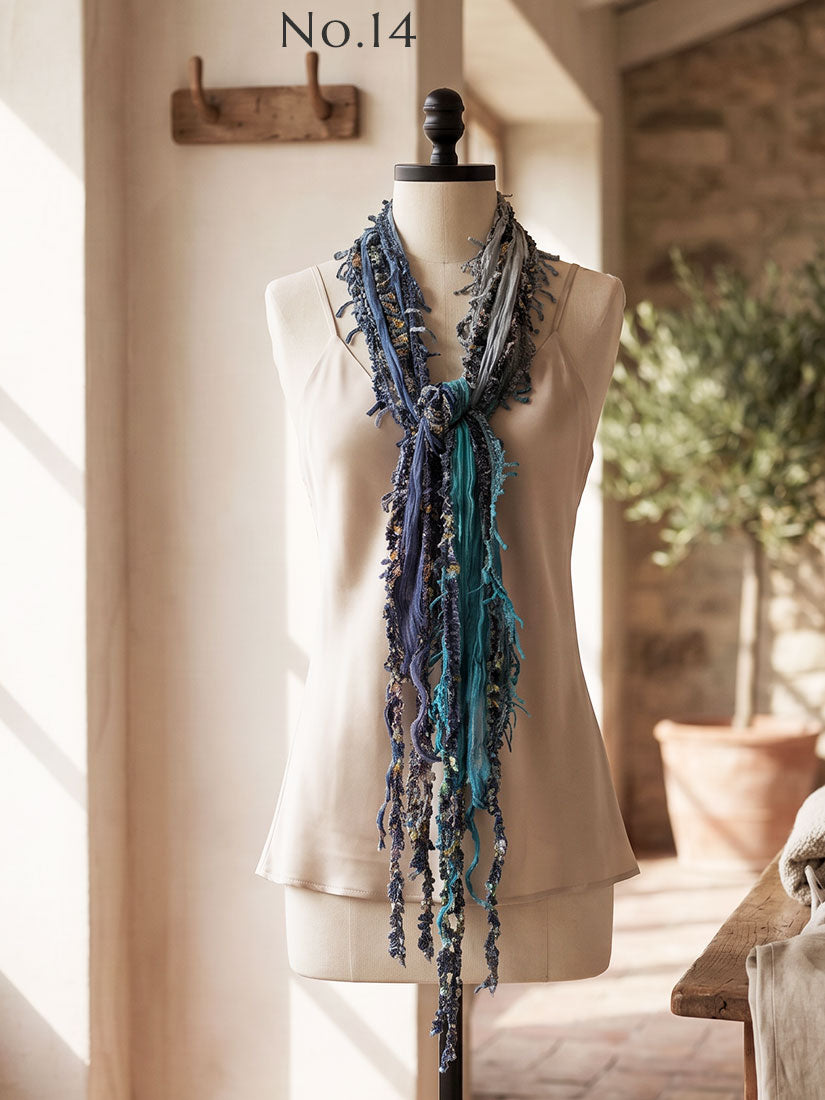 A long fringed scarf in shades of blue, teal, and gray with small white floral details by Monkey Mango, displayed on a cream-colored mannequin torso. The image is set in a warm, sunlit room with a rustic stone wall and potted plants in the background.