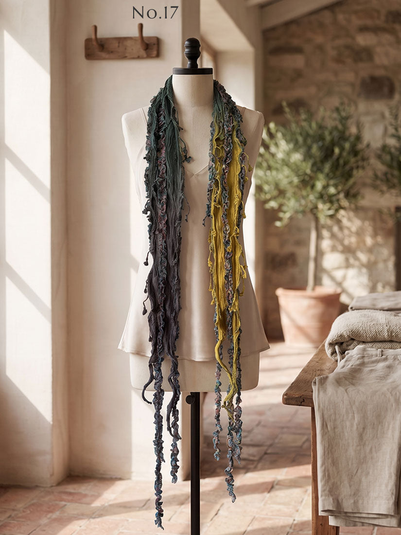 A long fringed scarf in muted gray-green and mustard yellow by monkey mango. draped over a cream mannequin torso in a rustic indoor setting with warm sunlight and stone walls.