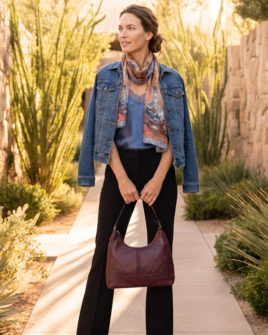 A woman stands on a sunlit walkway outdoors wearing a blue denim jacket, blue top, patterned scarf, black pants, and carrying a dark burgundy leather shoulder bag.