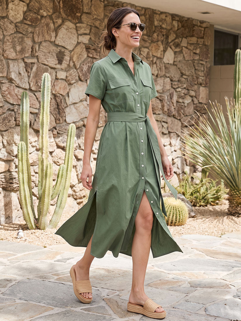 A woman wearing an olive green short-sleeve button-front midi dress walks outdoors on a stone patio in front of a stone wall with cacti.