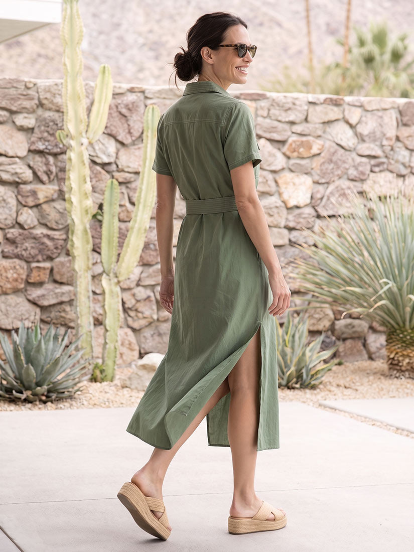 A woman wearing an olive green short-sleeve midi shirt dress with a side slit, walking outdoors on a paved path in front of a stone wall and desert plants.