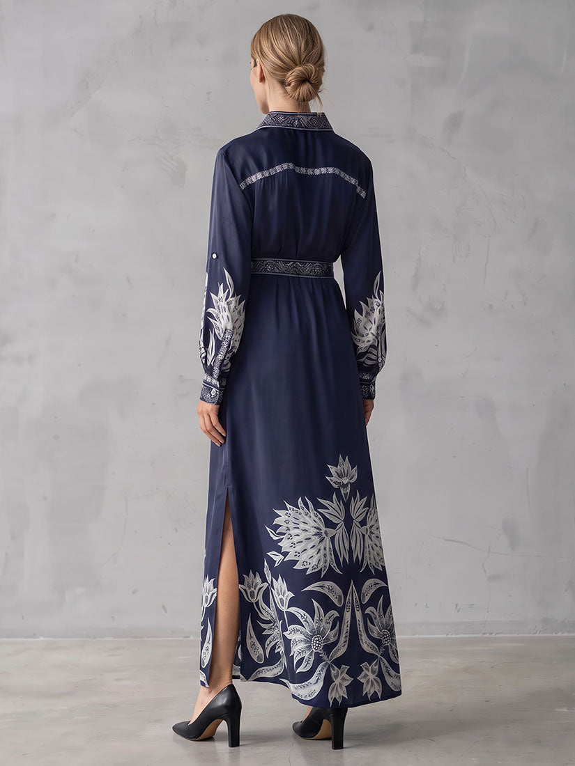 A woman viewed from the back wearing a long navy maxi dress with white botanical print and a matching dark belt, standing against a light gray textured wall.