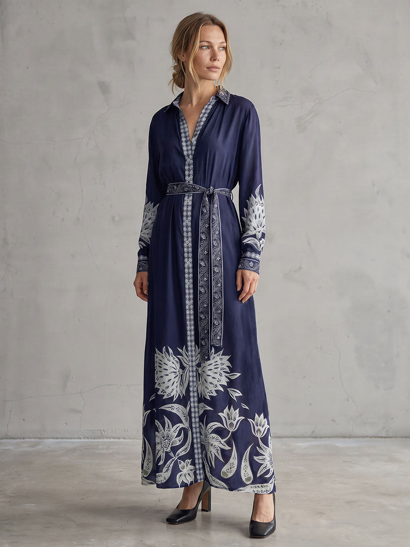 A woman models a navy blue maxi shirt dress with white floral prints and a matching patterned belt, standing against a textured gray concrete wall.