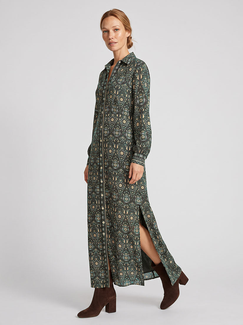 A woman modeling a long-sleeved button-front maxi dress with an intricate dark green and beige paisley pattern, featuring a high side slit and brown ankle boots, set against a plain light gray studio background.