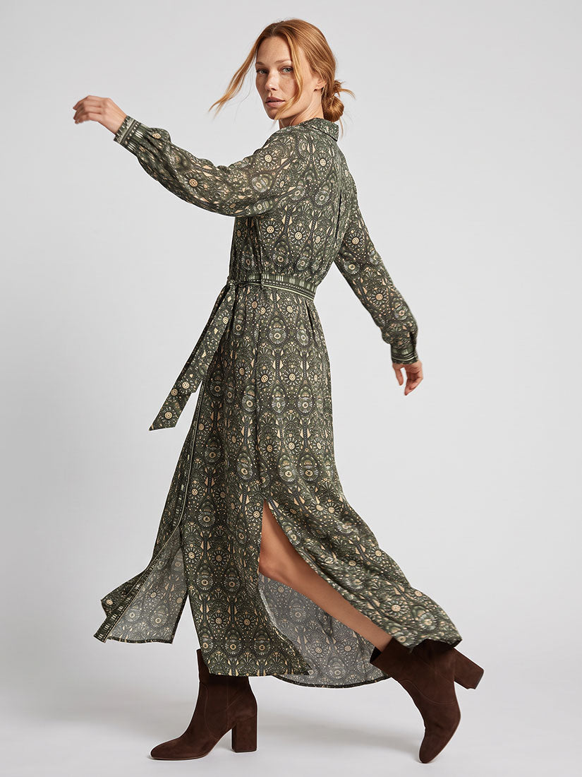 A woman modeling Omika emma  long-sleeved, high-neck maxi dress with a dark green and beige geometric pattern, featuring a tie waist and a high slit. She is wearing dark brown suede ankle boots against a light gray studio background.