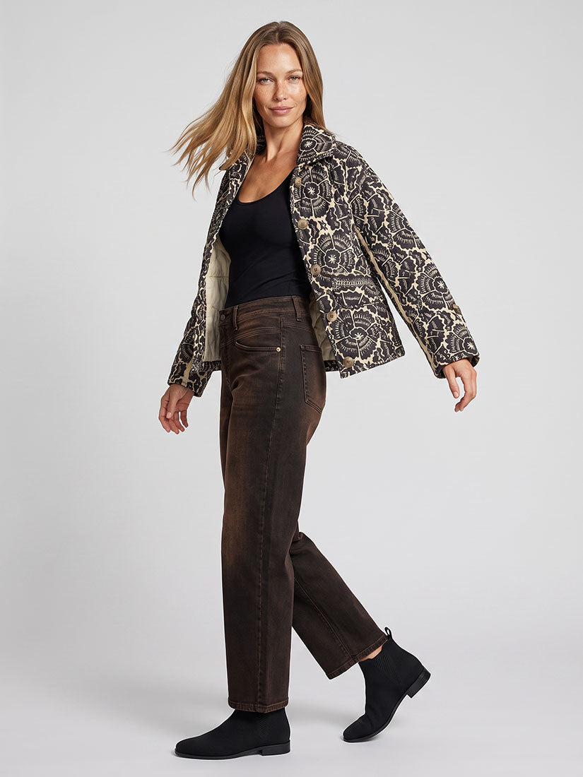 A woman modeling a cropped jacket with a black and beige floral pattern, worn over a black top and dark brown jeans, against a light gray studio background.