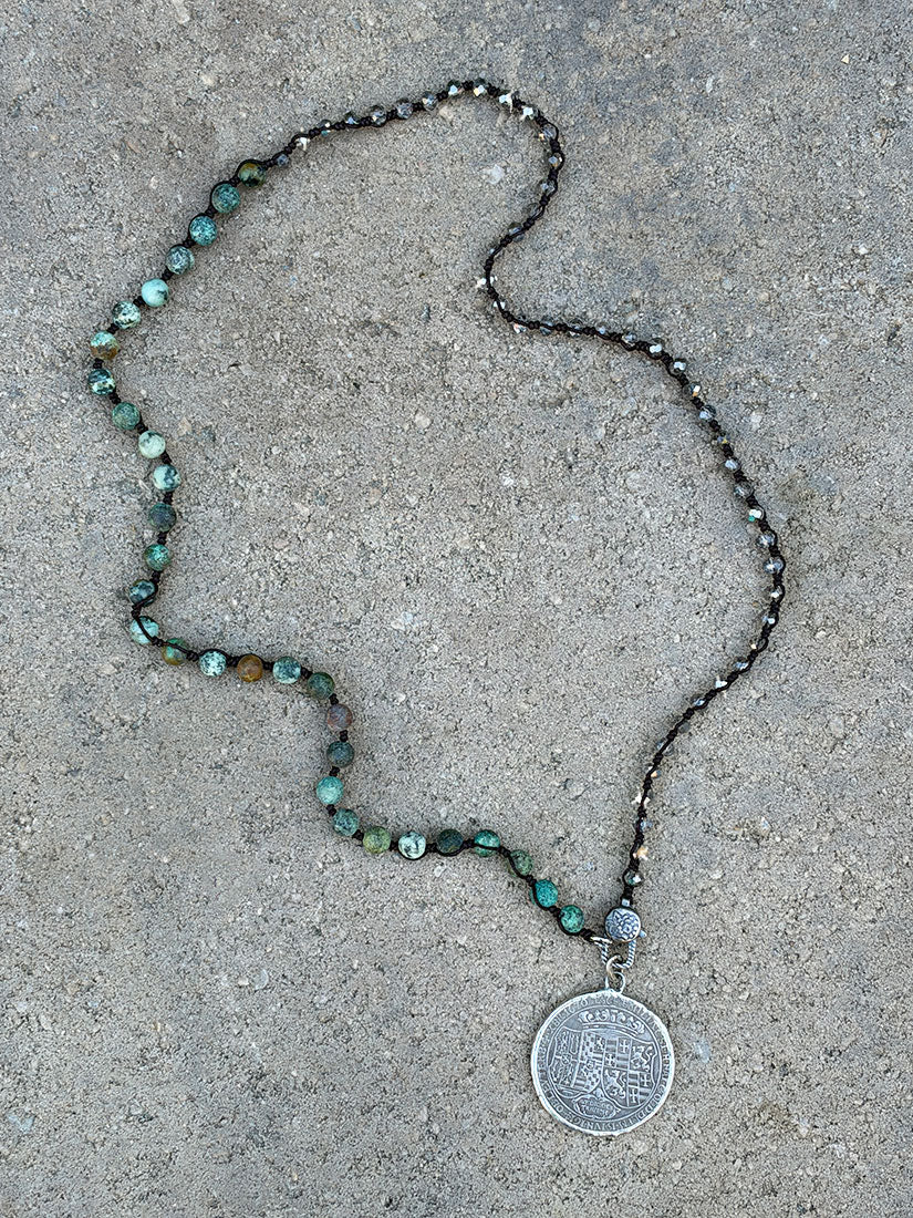 Hand knotted beaded necklace with green turquoise and smokey crystal beads from paula carvalho, featuring a round silver coin pendant, laid out on a textured gray concrete surface.
