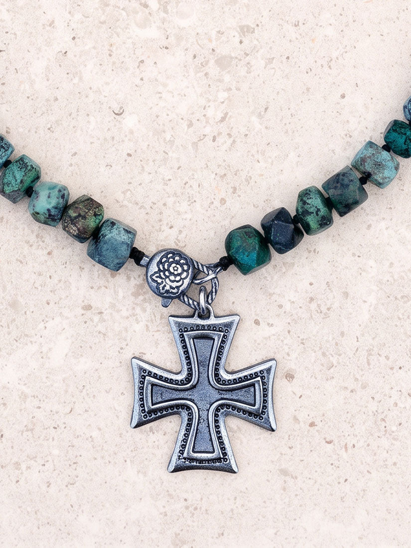 A close-up of a beaded necklace with faceted green and blue-green stones, a small silver-toned floral bead, and a large silver-toned cross pendant, laid on a light beige textured surface.