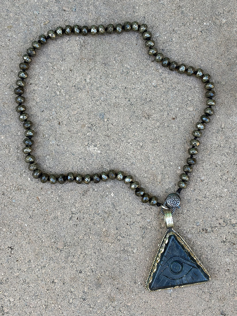 A beaded necklace with pyrite beads and a triangular carved dark blue agate pendant featuring an eye design, laid flat on a textured concrete surface.
