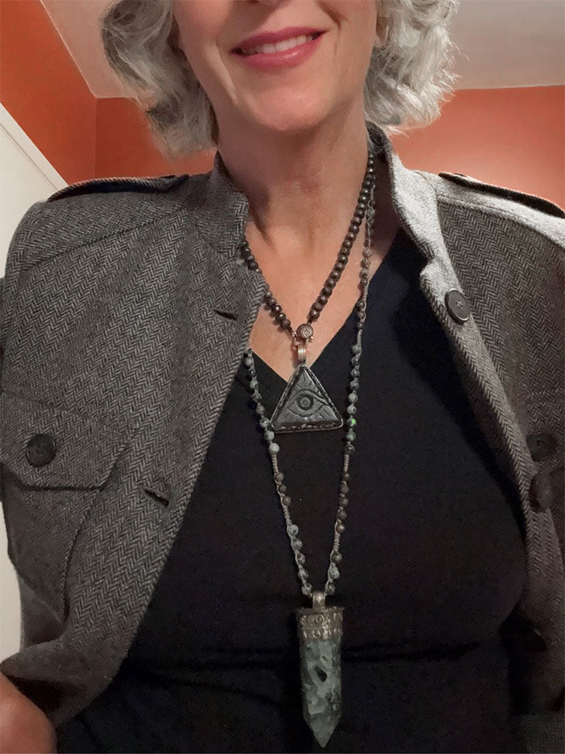 A close-up of a woman wearing a layered necklace set with a large triangular pendant necklace from paula carvalho and a long pointed stone pendant, styled with a gray blazer over a black top.