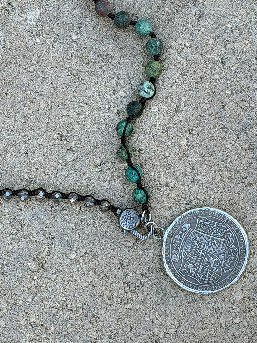 A close-up of a necklace laid on a textured concrete surface, featuring green and turquoise beads on a handknotted cord,  and a large round silver coin pendant.