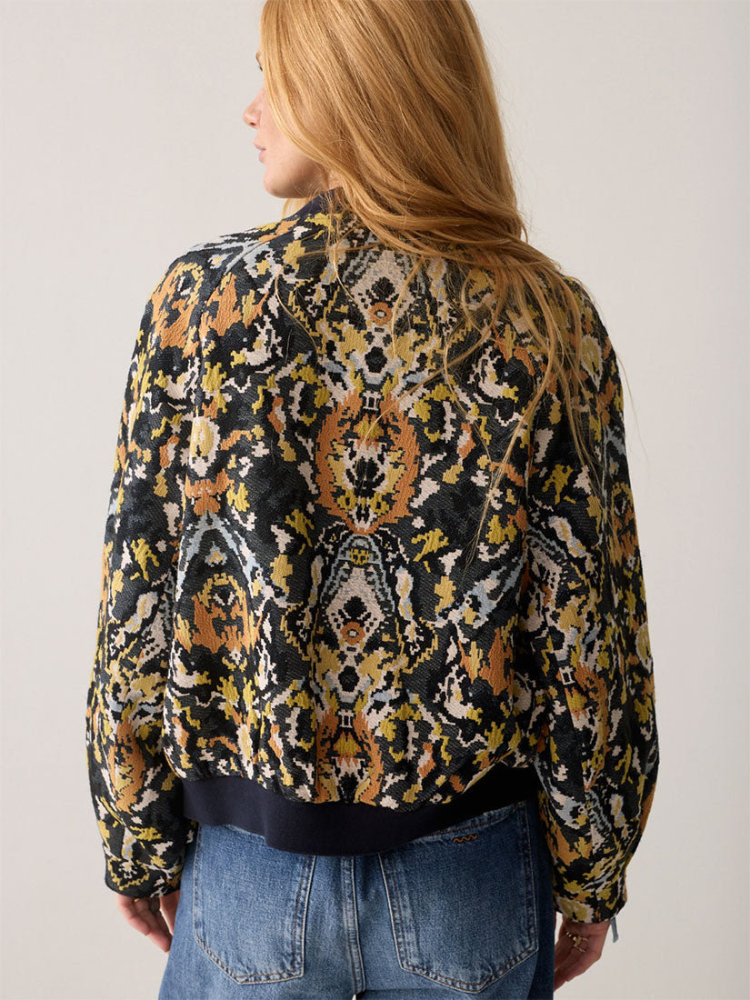 A woman shown from the back wearing a long-sleeve patterned jacket with a black base and intricate yellow, orange, and white ornamental print, paired with blue jeans against a plain light background.