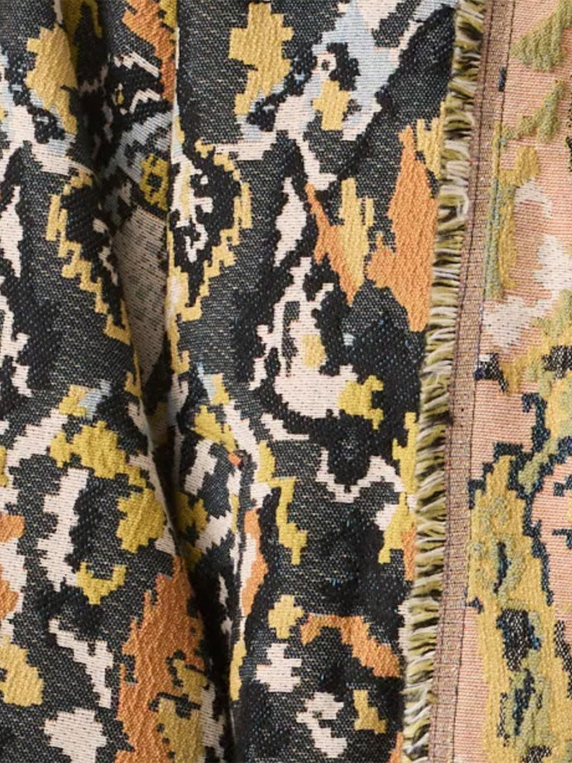 Close-up of a woven textile with an abstract geometric pattern in black, mustard yellow, peach, cream, and gray, with a fringed edge visible on the right.