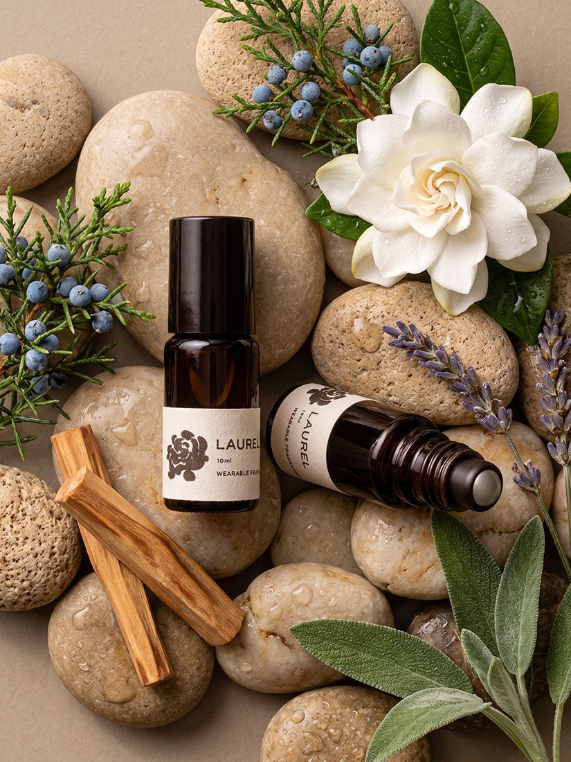 Two amber glass roller bottle bottles labeled LAUREL & ROSE essential oils, styled in a flat-lay with natural stones, lavender, sage, blueberries, and a white flower on a beige background.