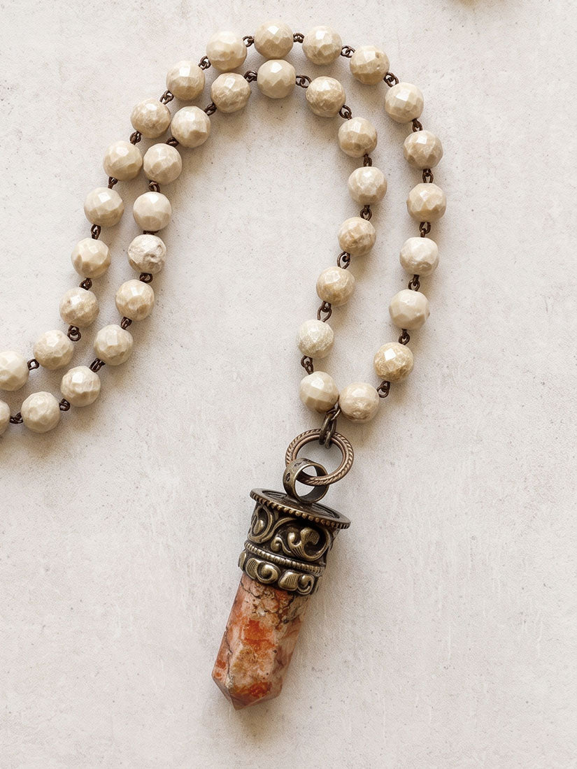 A beaded necklace with cream-colored faceted beads, dark metal spacer rings, and a pendant featuring a textured orange-brown stone capped with an ornate bronze-toned metal collar, displayed on a light beige textured background.